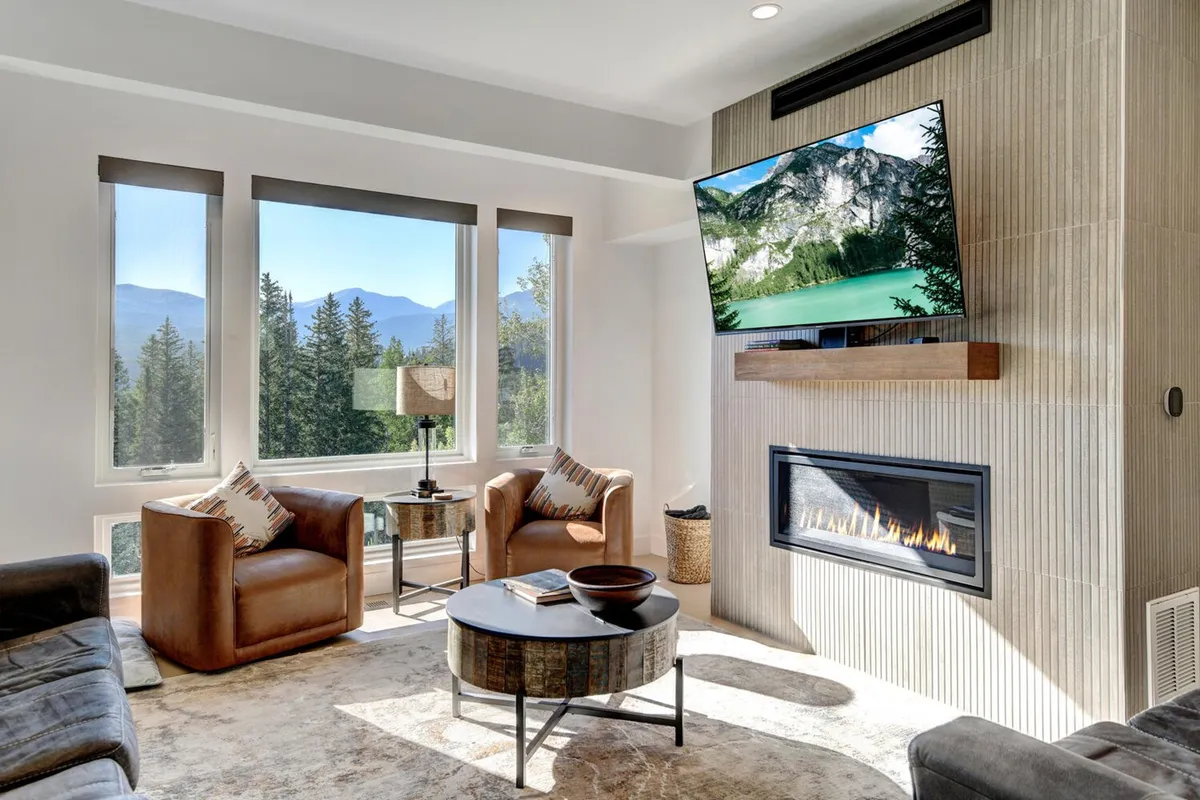 A cozy living room scene centers on a modern gas fireplace with a mounted HD television above, surrounded by comfortable sofas and large windows. The setting is ideal for socializing, movie nights, or relaxing after a day of adventure, with ample light and scenic views visible through the windows.