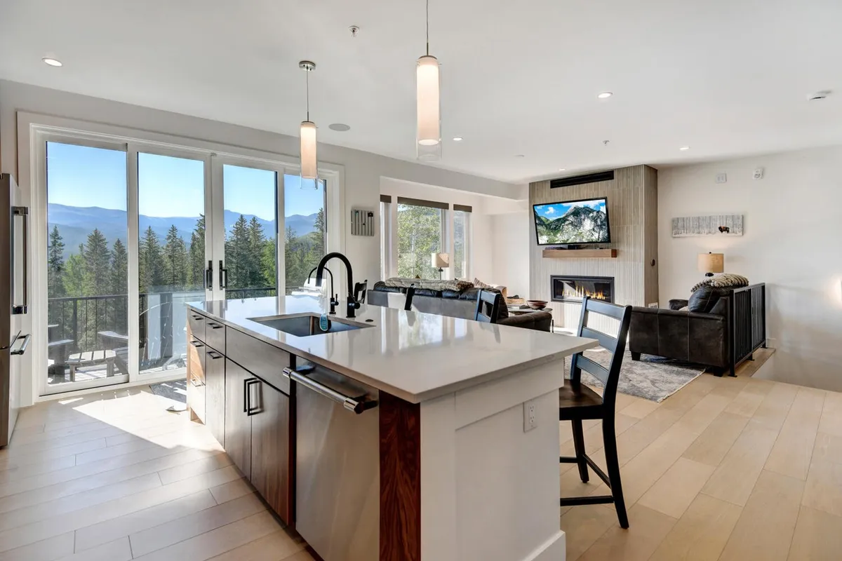 A bright, open interior showcases a kitchen island, a living area with a cozy fireplace, and expansive windows revealing stunning mountain views. The scene radiates warmth and comfort, hinting at evenings spent relaxing by the fire after a day of outdoor exploration.