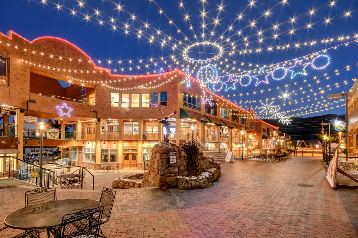 A warmly lit exterior scene featuring decorative string lights, a gently flowing stone fountain, and the facade of a building that suggests a welcoming downtown environment. The space is lively and inviting, perfect for evening strolls, dining, and social gatherings.