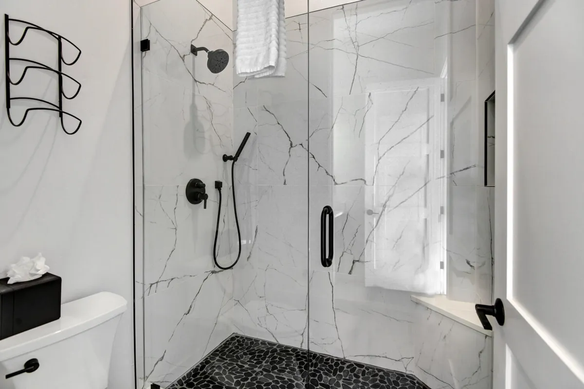 A modern, spa-inspired bathroom with a glass-enclosed shower, marble wall tiles, sleek black fixtures, and textured pebble floor tiles. The space is designed for soothing self-care, with light streaming in to enhance the tranquil vibe.
