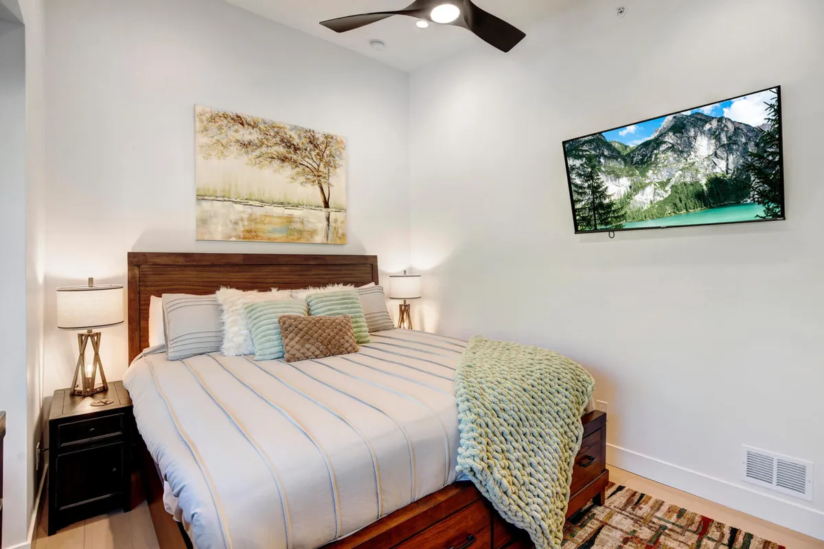 A tranquil bedroom featuring a plush bed adorned with decorative pillows, framed artwork above the headboard, and a wall-mounted HD television. Soft, natural light fills the space, creating a soothing atmosphere ideal for relaxation and unwinding after a day of adventure.