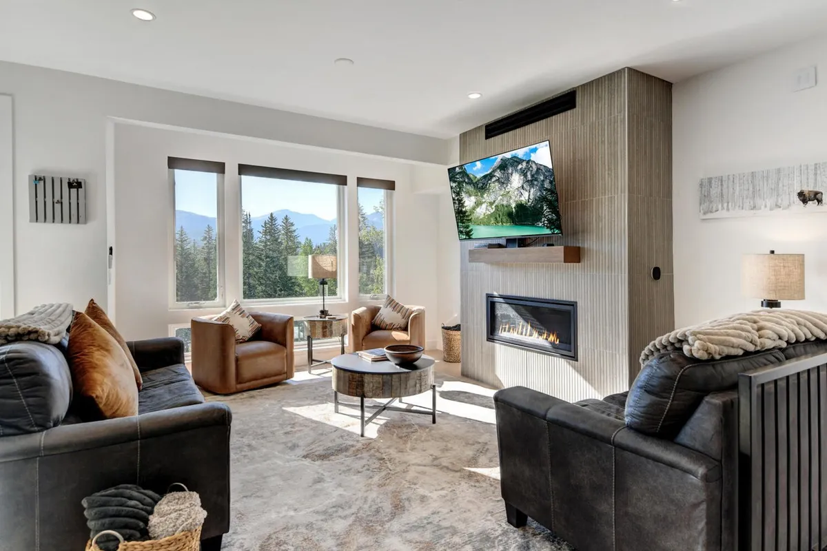 The image showcases a bright, modern living room with expansive windows framing breathtaking mountain views. A sleek fireplace anchors the space, surrounded by plush seating ideal for relaxing, socializing, or cozying up for movie nights. The room exudes warmth and comfort, with the natural scenery just beyond the glass inviting a sense of peace and escape.
