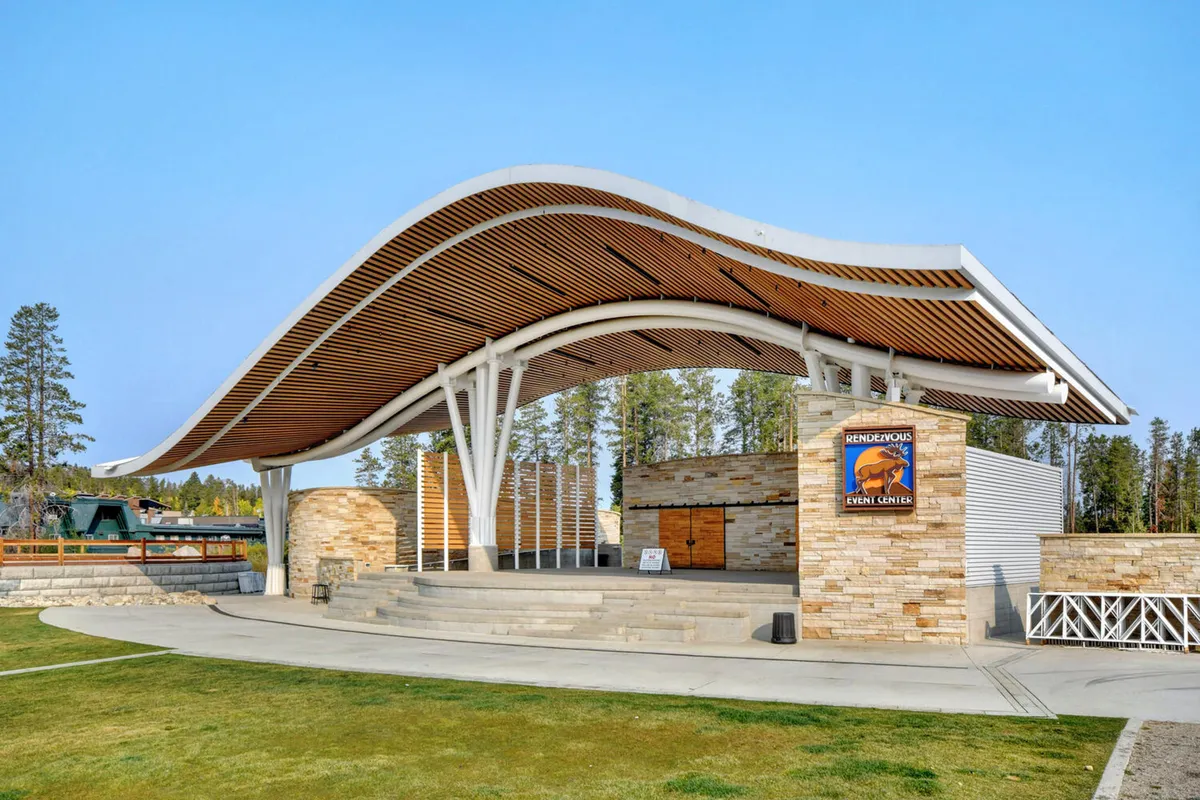 An exterior view of a modern event center with a distinctive curved roof, stone facade, and visible signage indicating its community function. The setting hints at concerts, festivals, and local gatherings under open skies.