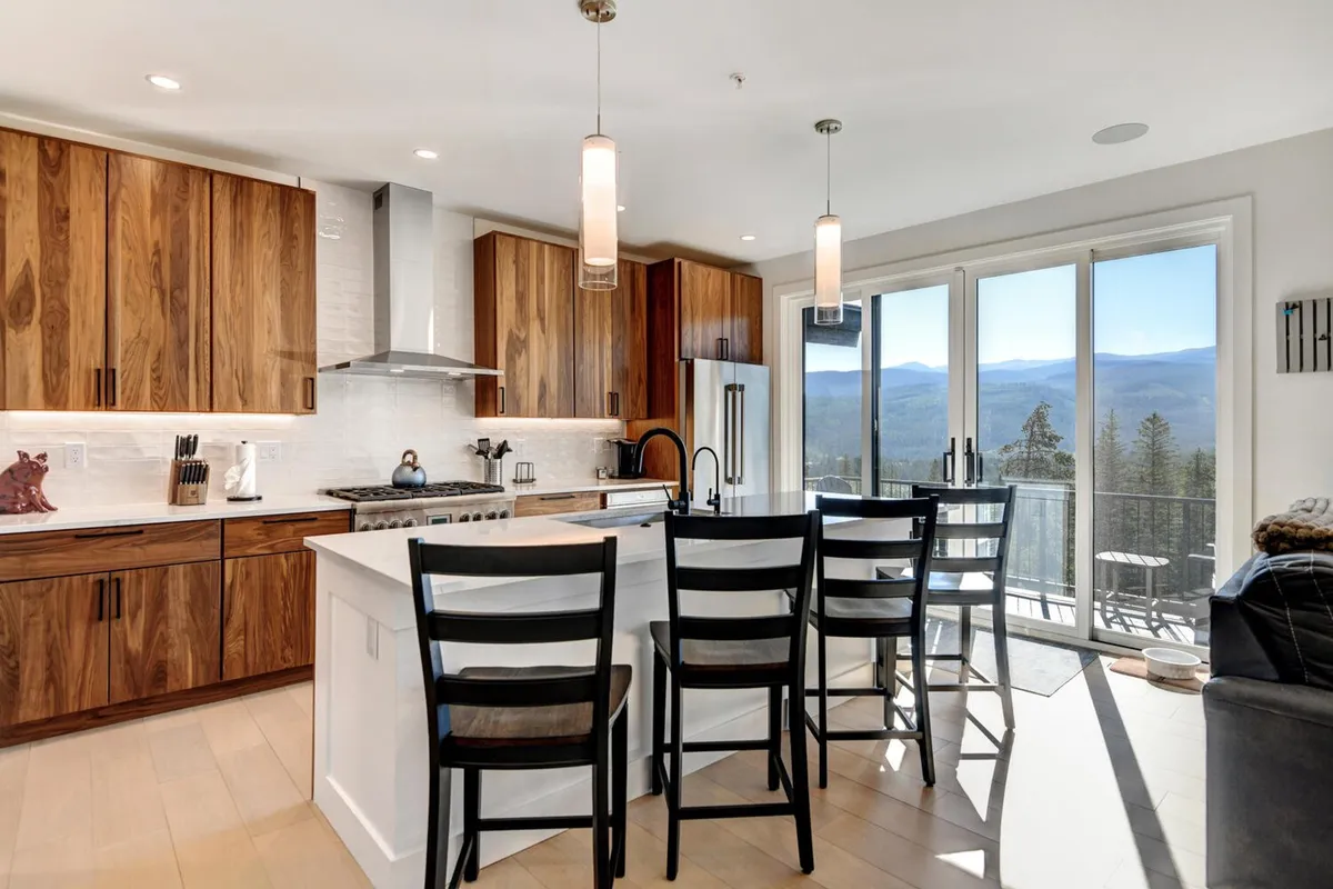 A warm, welcoming kitchen with a spacious island in the center, surrounded by sleek wood cabinetry and stainless steel appliances. Large windows frame breathtaking mountain views, and the space is set for cooking, dining, and entertaining. The island offers plenty of room for meal prep and gatherings, and the layout invites guests to participate in the culinary experience.