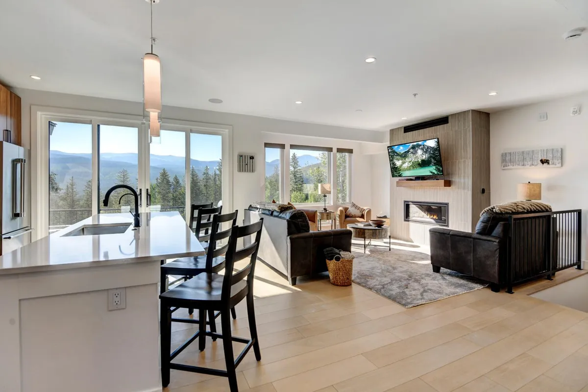 An expansive open-concept space seamlessly combines the living area, kitchen, and dining zone. Large windows flood the room with natural light and frame panoramic Rocky Mountain views. There’s a comfortable seating area with plush furnishings, a gas fireplace, a wall-mounted flat-screen TV, and a spacious kitchen island, all contributing to a welcoming and social environment.