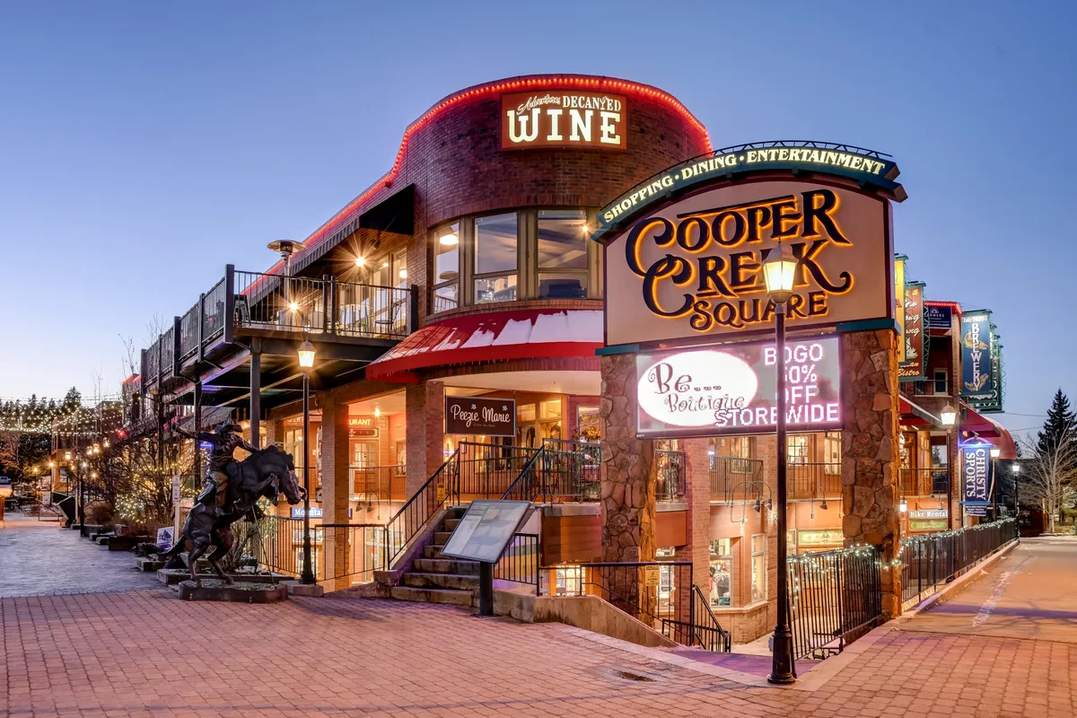 A bustling downtown scene featuring the Cooper Creek Square sign, inviting outdoor seating areas, a wine shop, and a modern sculpture. People enjoy shopping, dining, and gathering in a lively atmosphere with vibrant colors and clear weather.